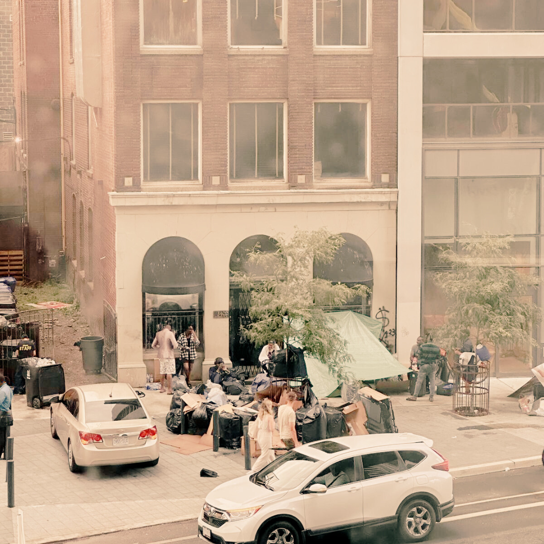 Image of migrants taking refuge on the sidewalk of a busy commercial street